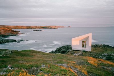 A small white building sits on the edge of a cliff, which has grass illuminated yellow by the setting sun. The cliff looks out onto a shoreline where waves crash into the rocks below.