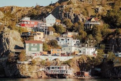 Colorful houses sit on a cliff beside the ocean. Between the buildings there are trees and long grass.