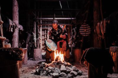 An indigenous person sits in the middle of a room holding a hand drum. They sit on a tree stump beside an open fire. The room is filled with wood, tools, and furs.