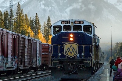 A train with a yellow mountain logo on the front sits still on track while people wait to board on the side platform. Behind the train stands a tall white mountain.