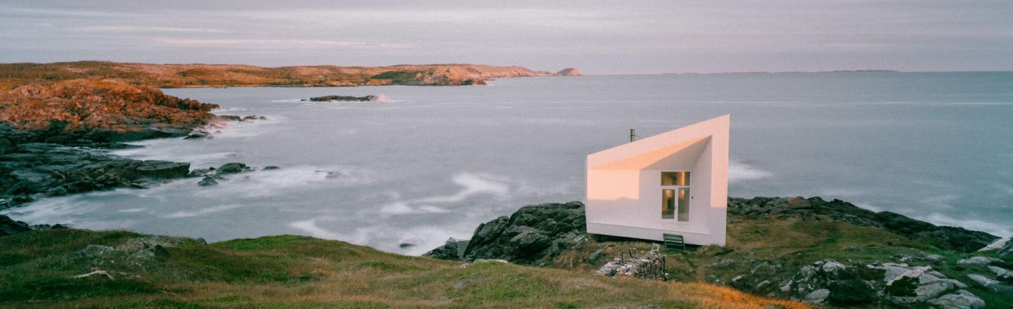 Un petit bâtiment blanc se tient sur le bord d’une falaise. Le soleil couchant projette une lueur dorée sur l’herbe. La falaise donne sur le littoral et des vagues s’écrasent sur les rochers au pied de la falaise.