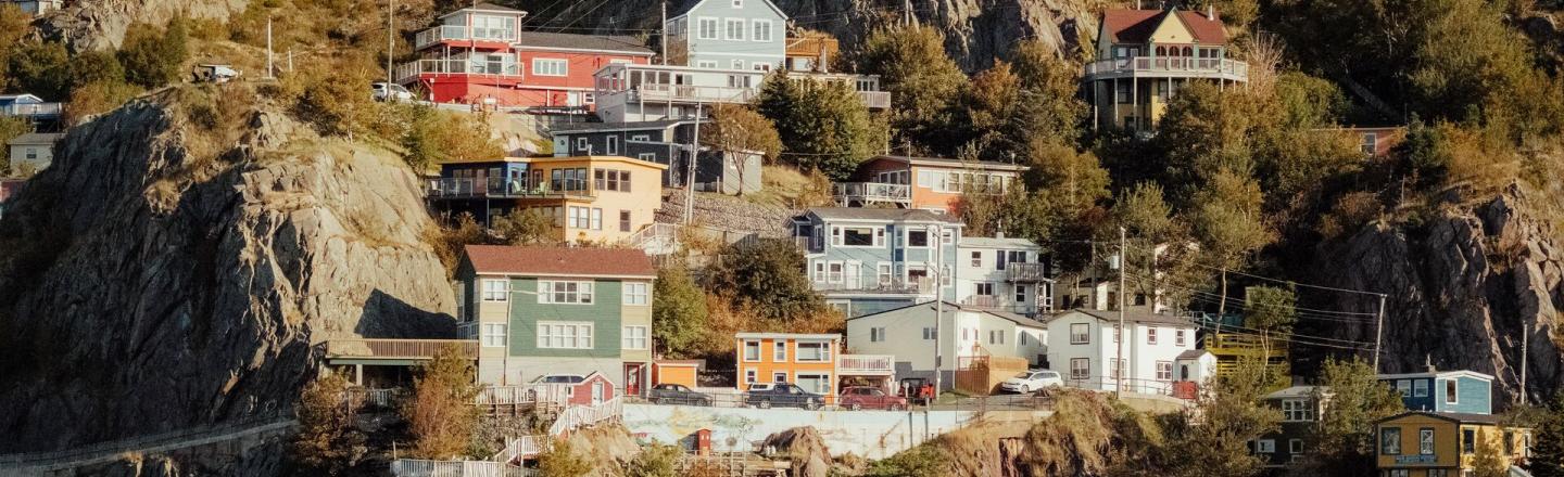 Colorful houses sit on a cliff beside the ocean. Between the buildings there are trees and long grass.