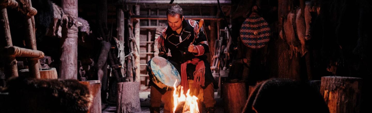 An indigenous person sits in the middle of a room holding a hand drum. They sit on a tree stump beside an open fire. The room is filled with wood, tools, and furs.