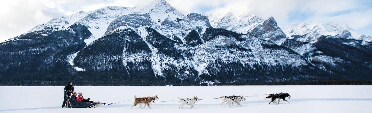 A team of eight dogs pull a dog sled across the snow. Two people sit in the front of the sled while a third stands behind. A towering white-capped mountain stands in the background against a partially cloudy sky.