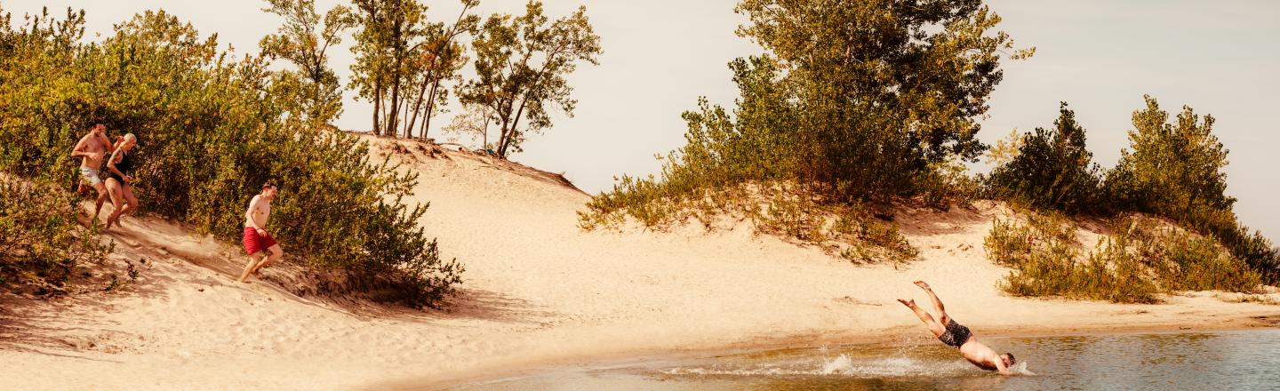 Trois personnes en maillot de bain courent sur une dune de sable aux abords d’un lac. Une quatrième personne, à l'avant, plonge dans l'eau. Le sable autour du lac est bordé d'arbres et d'arbustes verts.Trois personnes en maillot de bain courent sur une dune de sable aux abords d’un lac. Une quatrième personne, à l'avant, plonge dans l'eau. Le sable autour du lac est bordé d'arbres et d'arbustes verts.